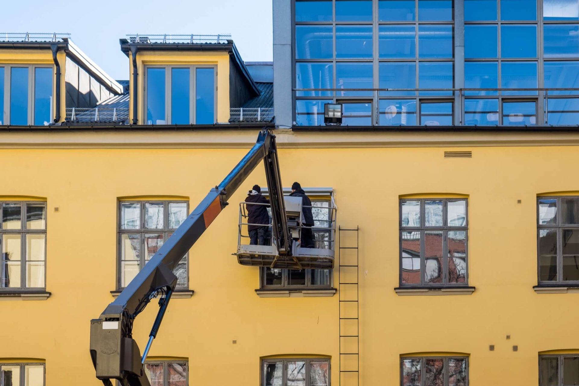 Två arbetare på en skylift engagerar sig i fasadarbete på en gul byggnad som är under renovering